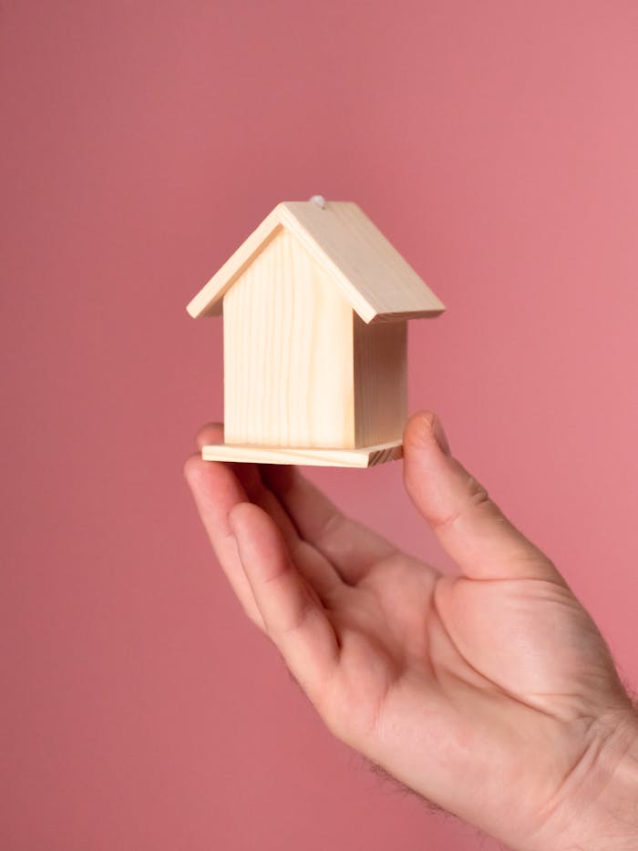 A close-up of a hand holding a small wooden house model against a pink background, symbolizing property and investment.